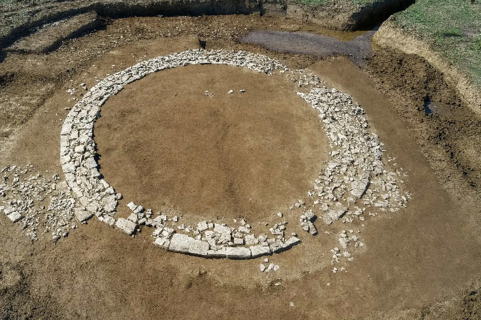 Umjetnička rekonstrukcija arheološkog nalazišta u Bavarskoj koja prikazuje kameni kružni temelj rimskog tumulusa (grobnog humka) otkrivenog u blizini mjesta Wolkertshofen. Prikaz temelji se na stvarnim fotografijama iskopavanja te vjerno dočarava izgled strukture i okolnog tla tijekom istraživanja.