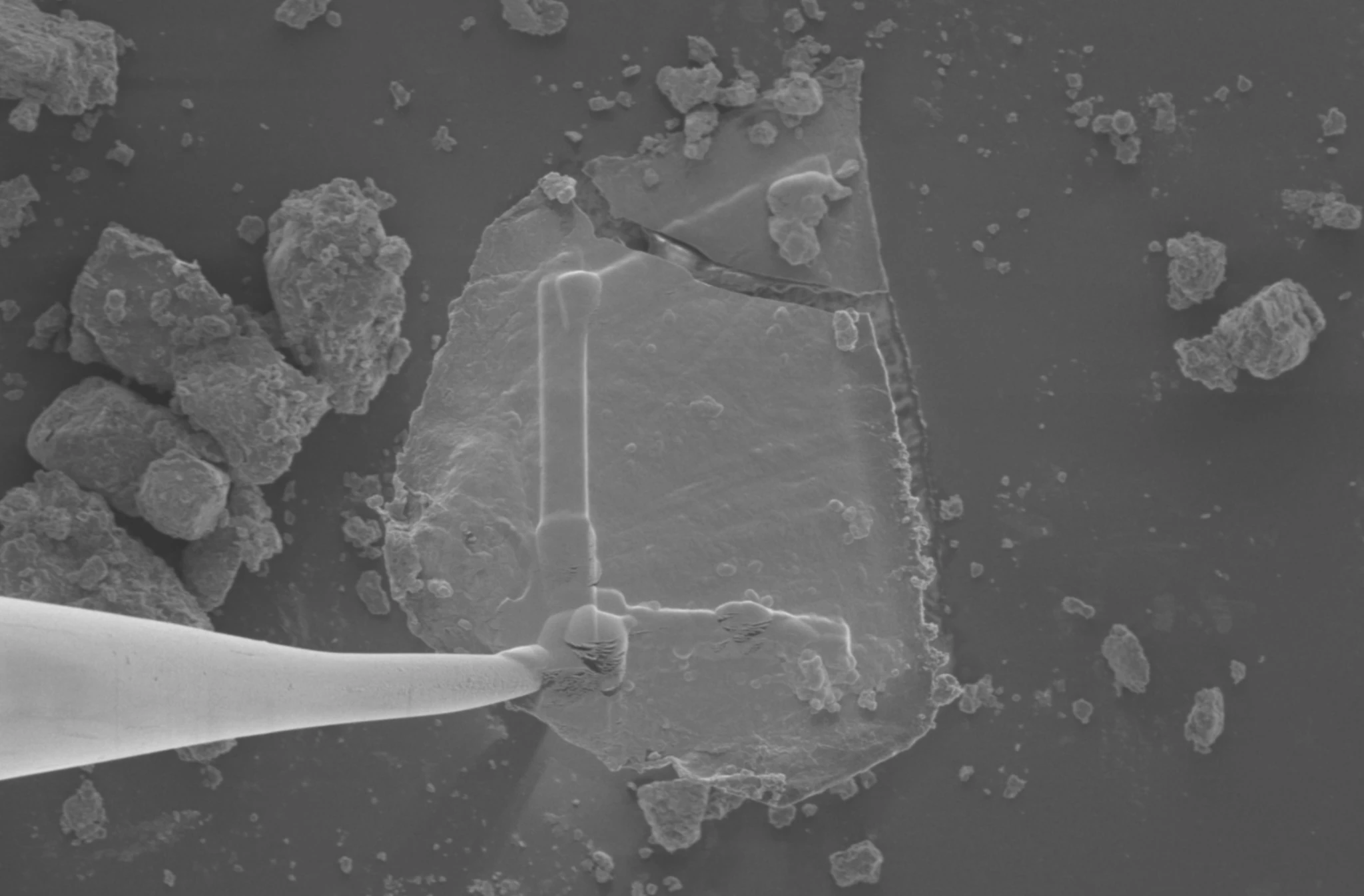 Mikroskopska čestica asteroida Bennu, koju je na Zemlju donijela NASA-ina misija OSISRIS-REx, nalazi se pod transmisijskim elektronskim mikroskopom. Kako bi fragment mogli pomaknuti na daljnju analizu, istraživači su ga najprije ojačali tankim trakama platine (oblik slova "L" na površini čestice), a zatim su na njega zavarili volframovu mikroiglu. Fragment asteroida širok je oko 30 mikrometara, usporedivo s veličinom sitnog zrnca prašine. NASA/University of California, Berkeley