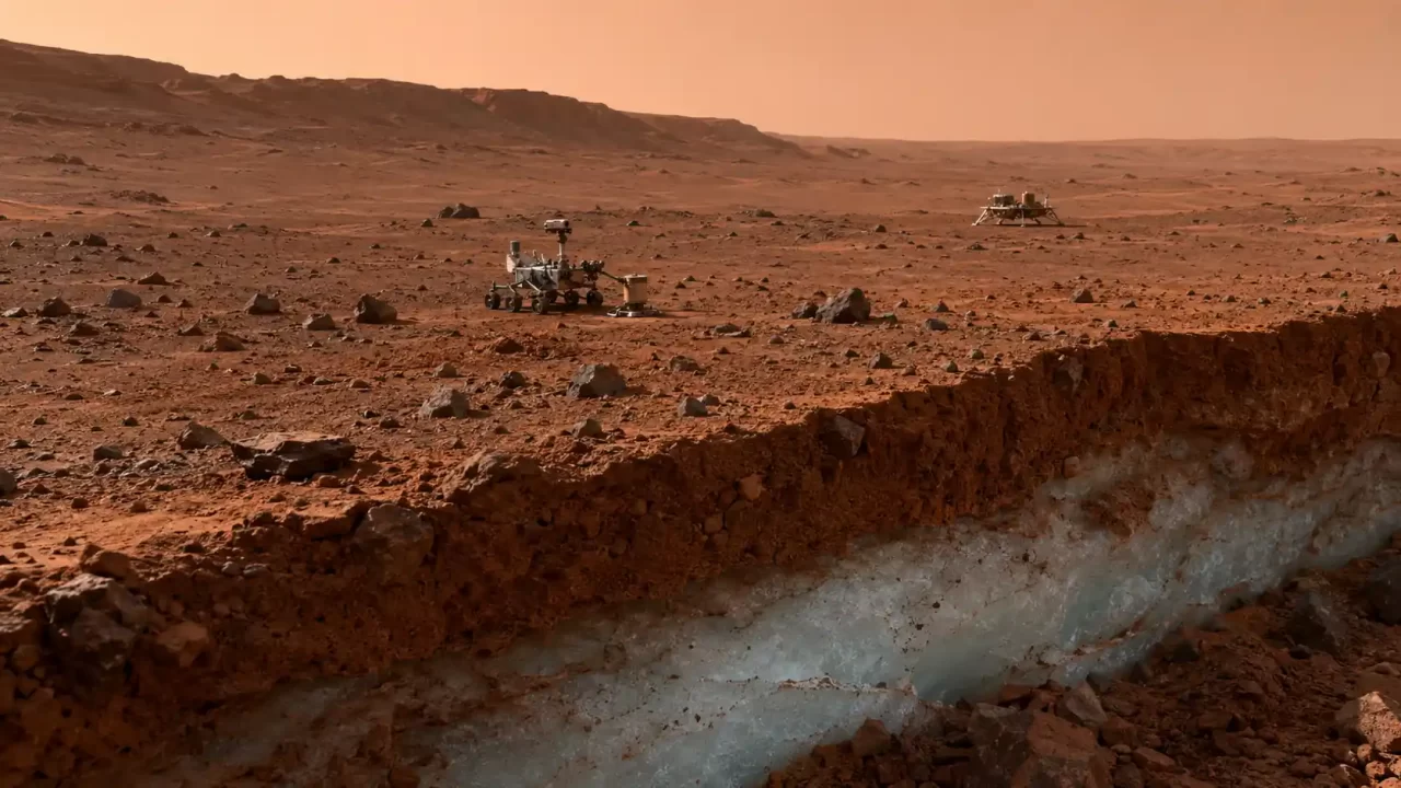 Marsovski rover i mali lender na kamenitoj površini Marsa, iznad presjeka tla koji otkriva sloj vodenog leda ispod crvenkastog regolita.