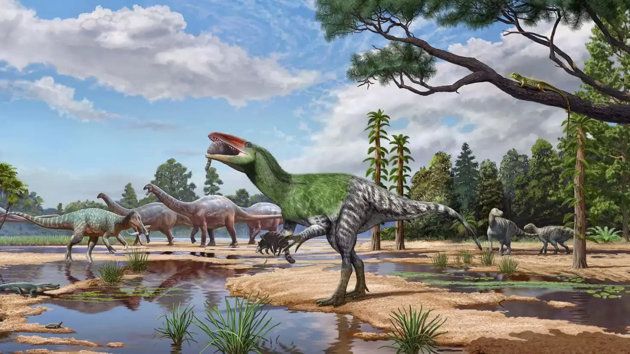 Pernati dinosaur lovi male sisavce na obali, uz velike dinosaure i raslinje u pozadini.