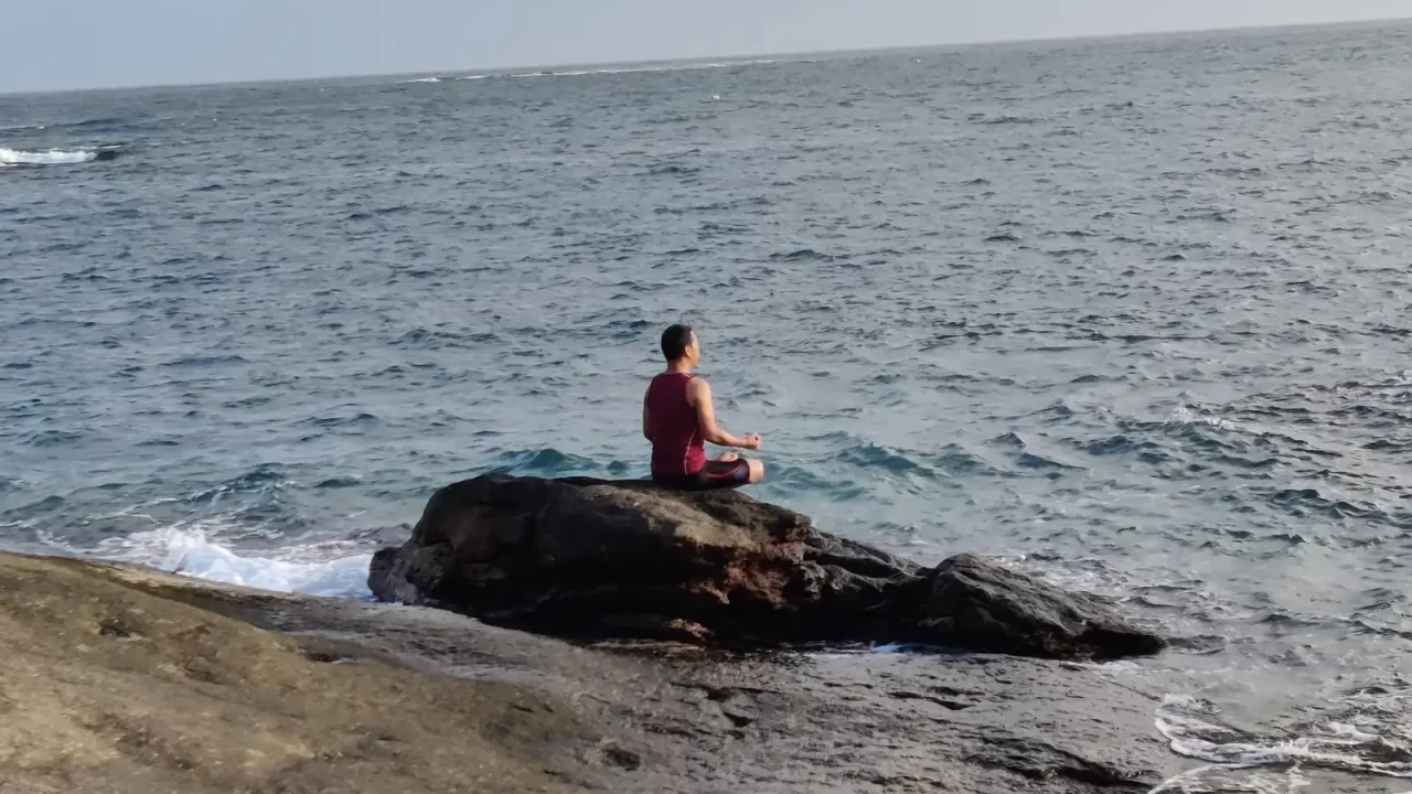 Osoba sjedi prekriženih nogu na stijeni uz more i meditira.