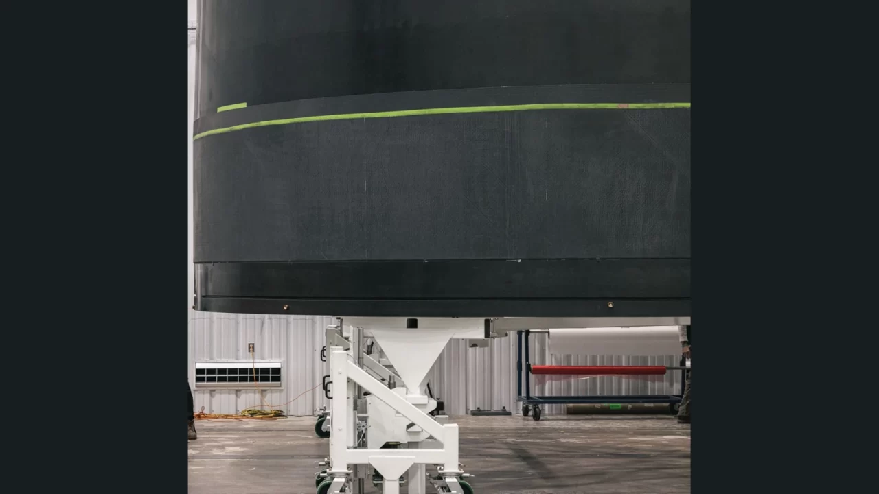 Velika tamna cilindrična kompozitna konstrukcija rakete Neutron na bijelom postolju u proizvodnoj hali.