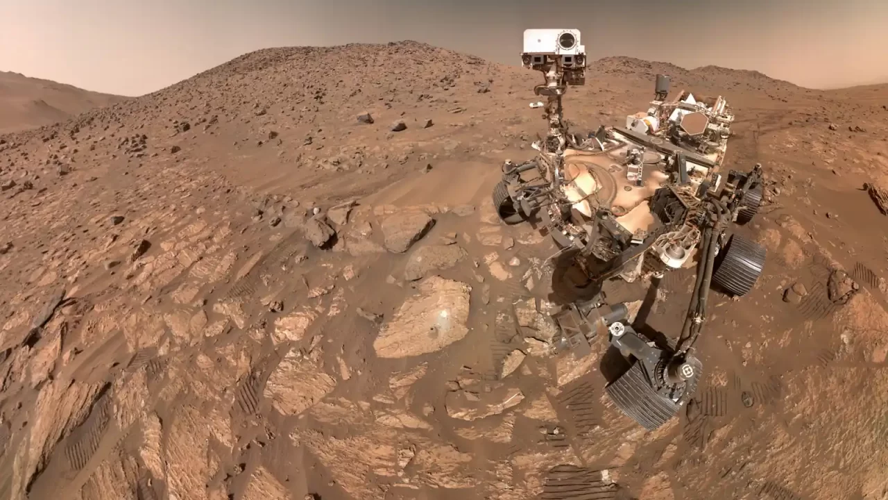 Rover Perseverance na kamenitom marsovskom tlu s tragovima kotača i brežuljcima u pozadini.